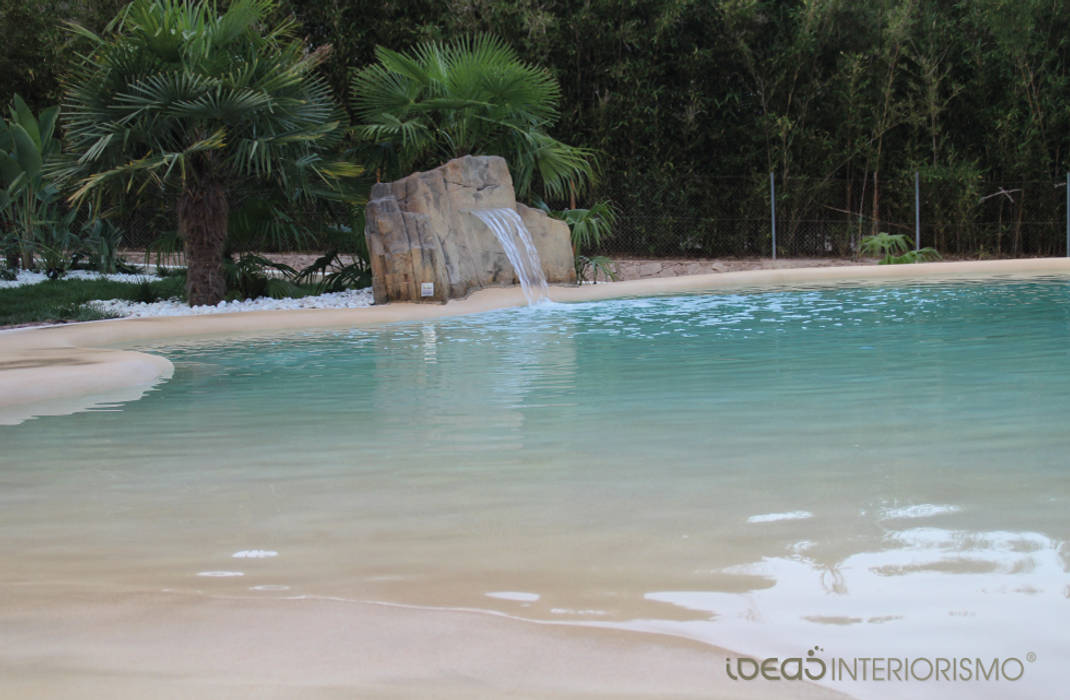 Paisajismo en un jardín con una piscina de arena. Ideas Interiorismo Exclusivo, SLU Piscinas de estilo mediterráneo Agua,Planta,Árbol,Azur,Paisaje natural,Accidentes geográficos costeros y oceánicos,Corriente de agua,arecales,Lago,Banco