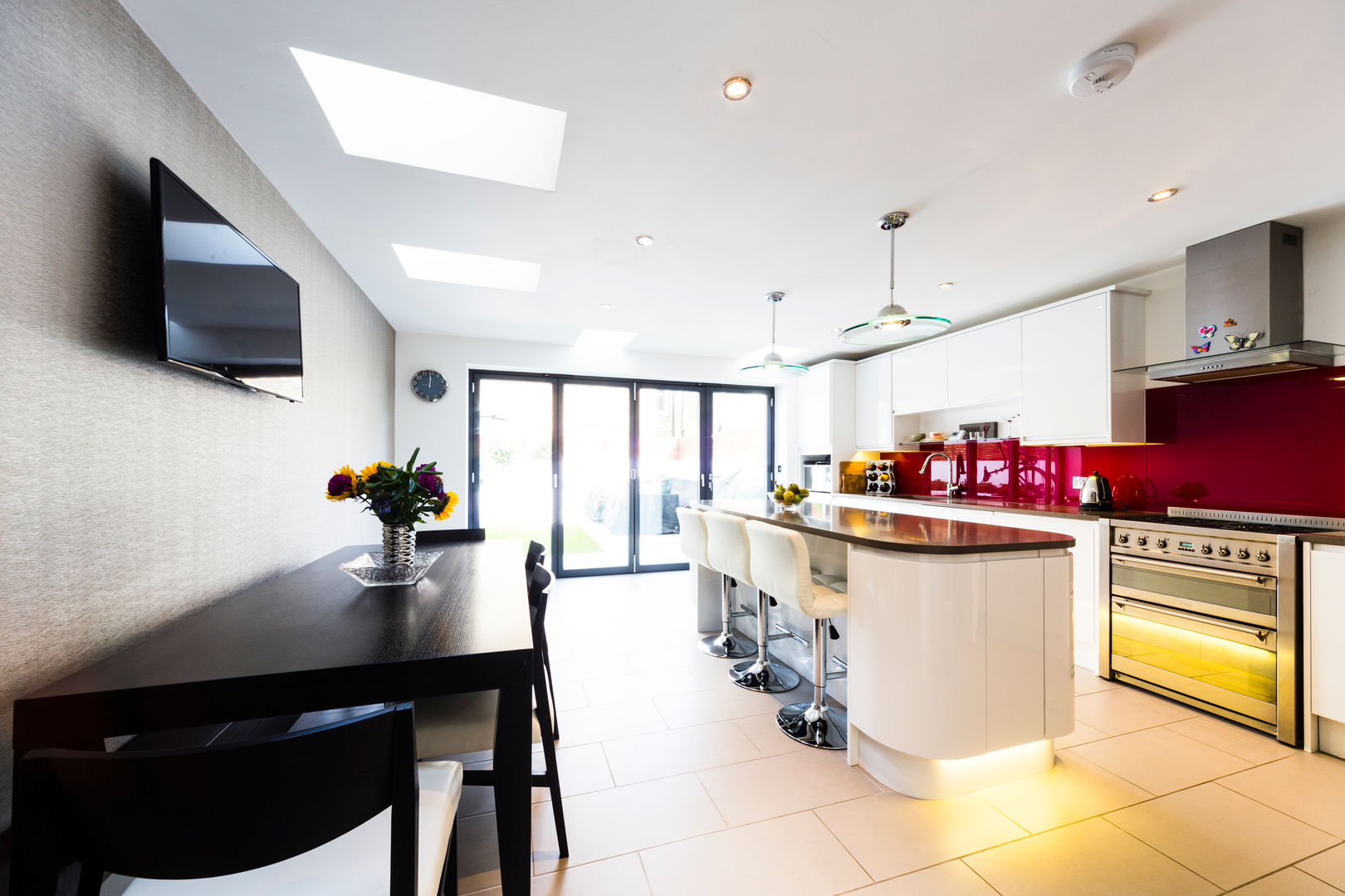 White kitchen with red splashback, modern kitchen pendants, bifold doors, black dining table and chairs Affleck Property Services Cocinas de estilo moderno